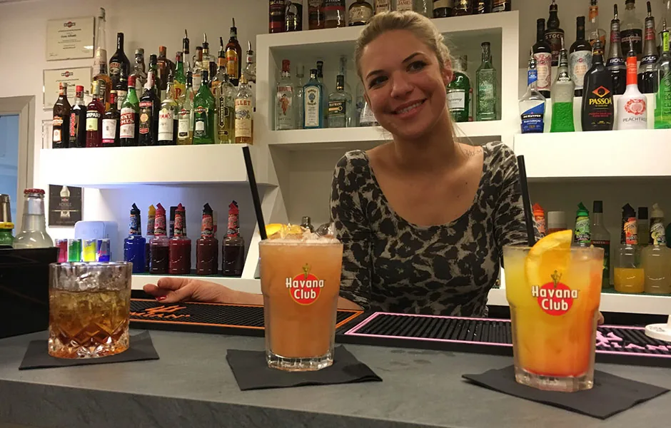 Una corsista sorride dietro al bancone con tre cocktail appena preparati